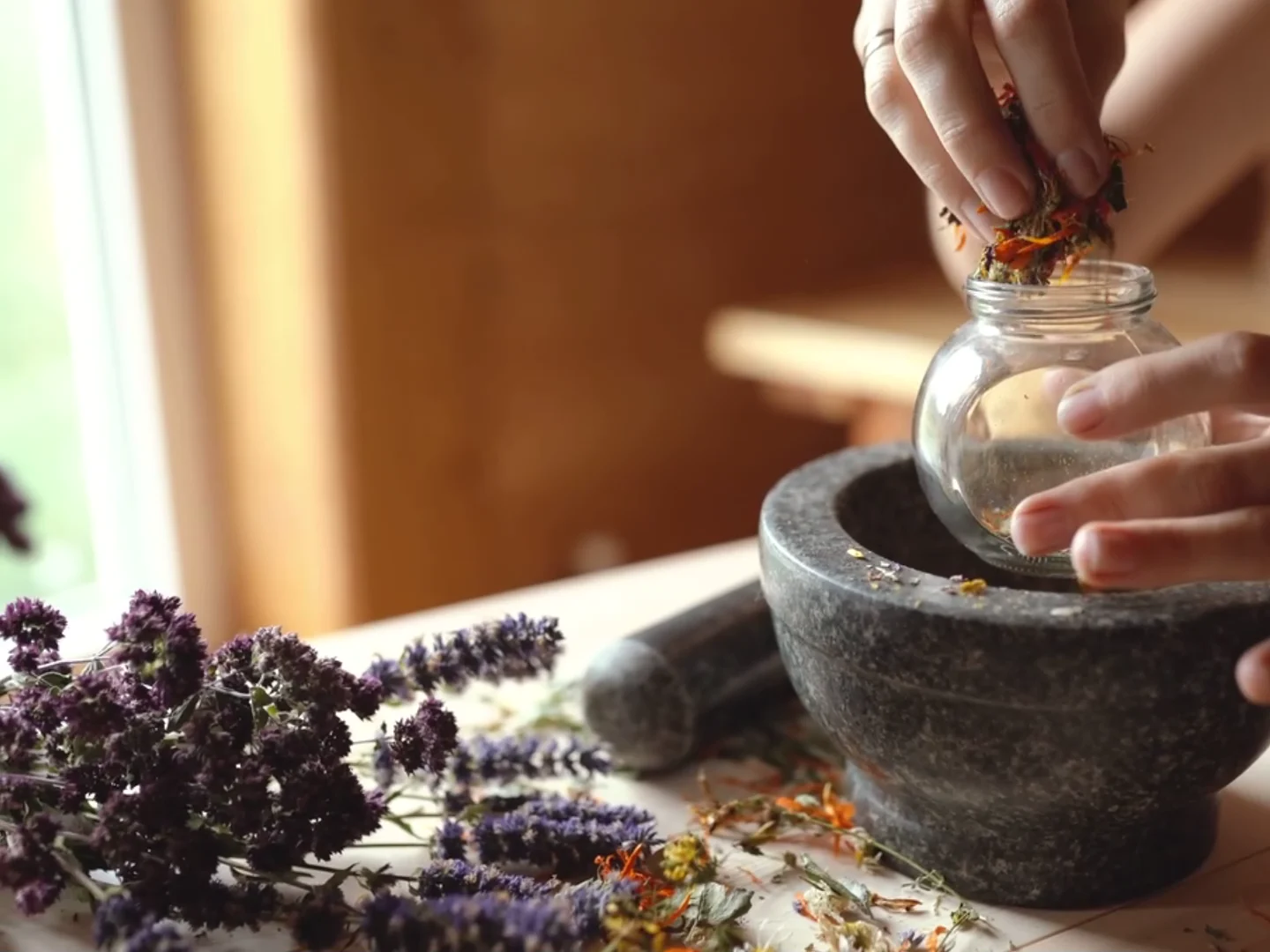 Hands preparing dried herbs beside a stone mortar and glass jar