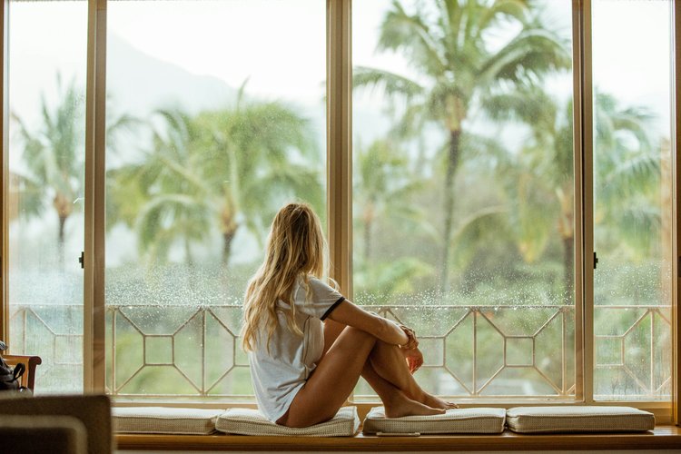 A woman sitting quietly beside a large window overlooking palm trees