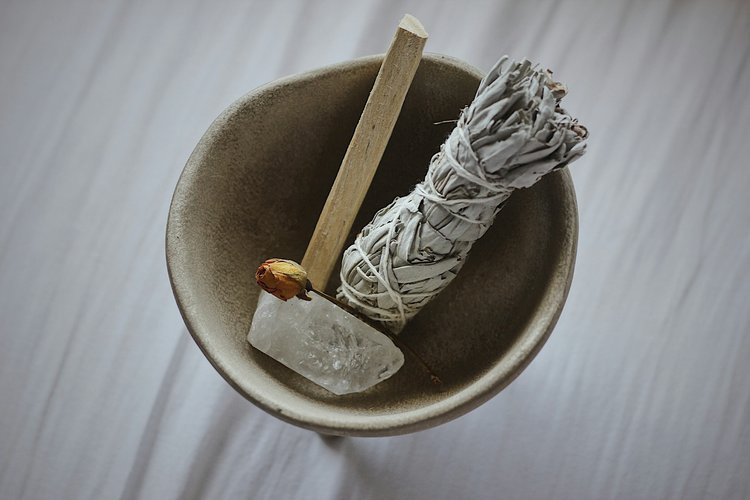 A ceramic bowl holding a dried sage bundle, a wooden stick, and a clear crystal