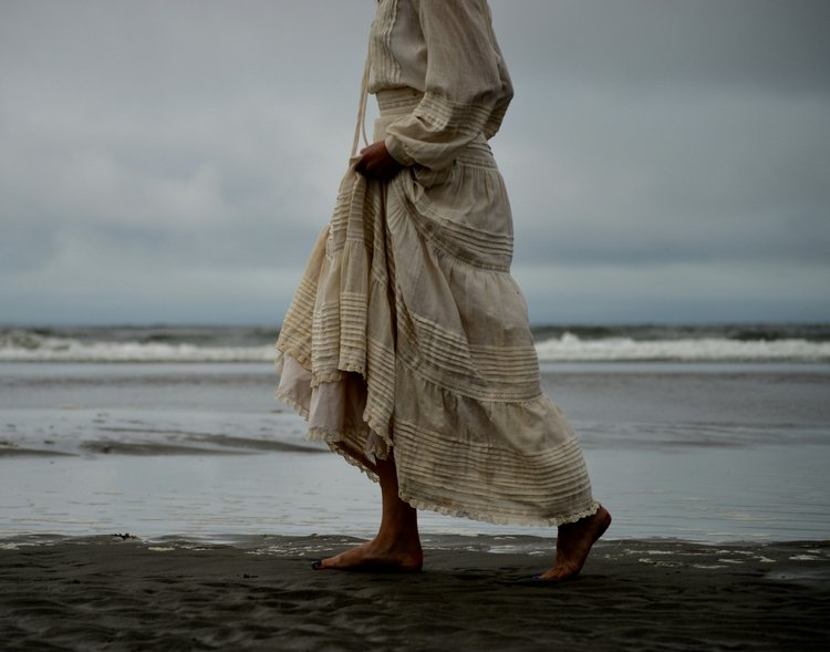 A woman in a flowing cream dress walking barefoot along the shore